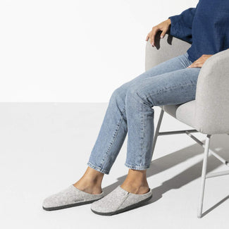 A light gray felt slip-on clog shoe with a dark gray textured sole and a small metal logo button on the side, shown against a plain white background.