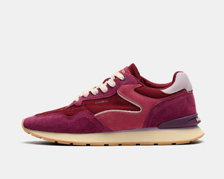 A low-top sneaker in shades of burgundy and magenta suede, featuring cream laces and a beige midsole, shown from a side profile on a light gray background.