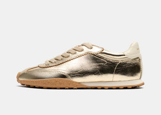 A metallic gold low-top sneaker with matching gold laces and a tan rubber sole, shown in profile against a light gray background.