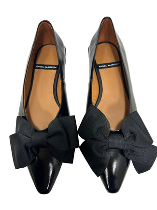 A pair of black leather low-heel flats with bow details on the toe, shown from a side profile against a plain light gray background.