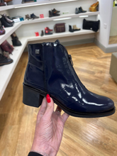 Load image into Gallery viewer, A navy blue patent leather ankle boot with a chunky black block heel, held by a hand with red nail polish in a shoe store.
