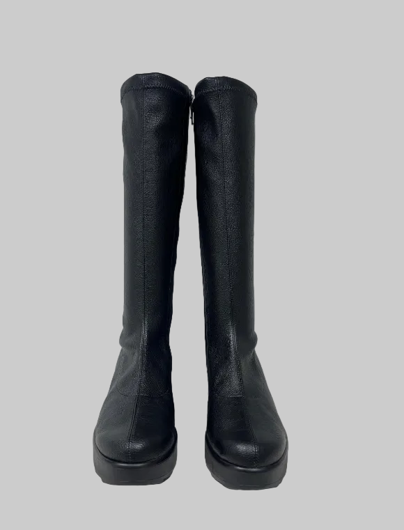 A pair of black knee-high leather boots with a chunky platform sole, standing upright against a plain light gray wall.
