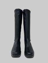 Load image into Gallery viewer, A pair of black knee-high leather boots with a chunky platform sole, standing upright against a plain light gray wall.
