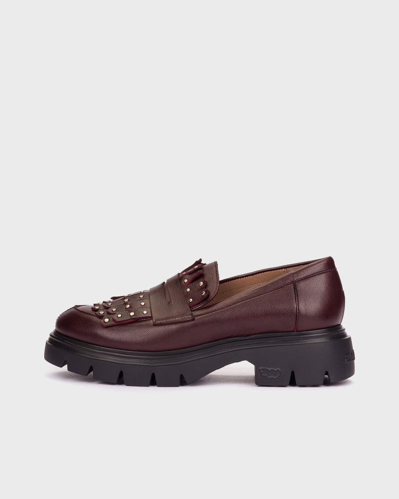 A single dark burgundy leather loafer with a chunky black lug sole and metallic studs on the strap, shown from a side profile against a light gray background.