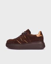 Load image into Gallery viewer, A dark brown suede low-top sneaker with a chunky ribbed platform sole, black laces, and metallic bronze accents on the heel and side, shown against a light gray background.
