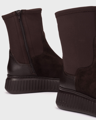 A dark brown ankle boot with a textured elastic side panel, a glossy leather toe cap, and a chunky ribbed platform sole, shown from a side profile on a light gray background.
