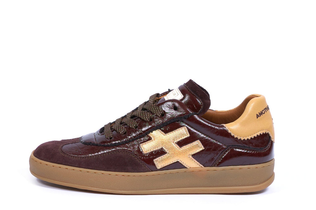 A low-top sneaker in dark brown leather with a tan cross logo on the side, a tan heel tab, and a tan gum sole, shown against a plain white background.