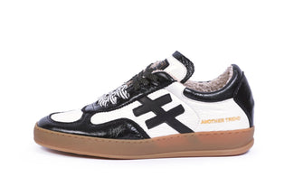 A low-top sneaker in black and white leather with a gum sole, featuring a large black side stripe and gold 'ANOTHER TRIO' branding on the heel.