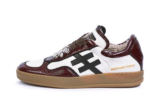 A low-top sneaker by Another Trend featuring dark brown and white leather panels, a black geometric side logo, brown laces, and a tan gum sole.