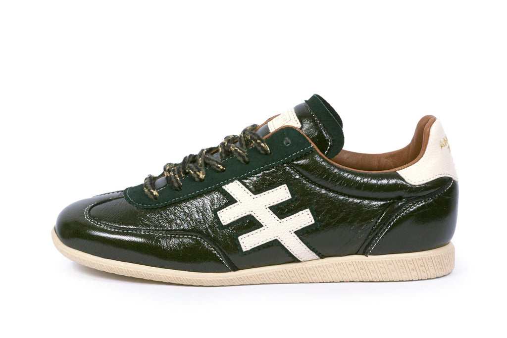 A dark green leather sneaker with a prominent white stylized 'X' logo on the side, dark green laces, and a beige sole, shown against a plain white background.