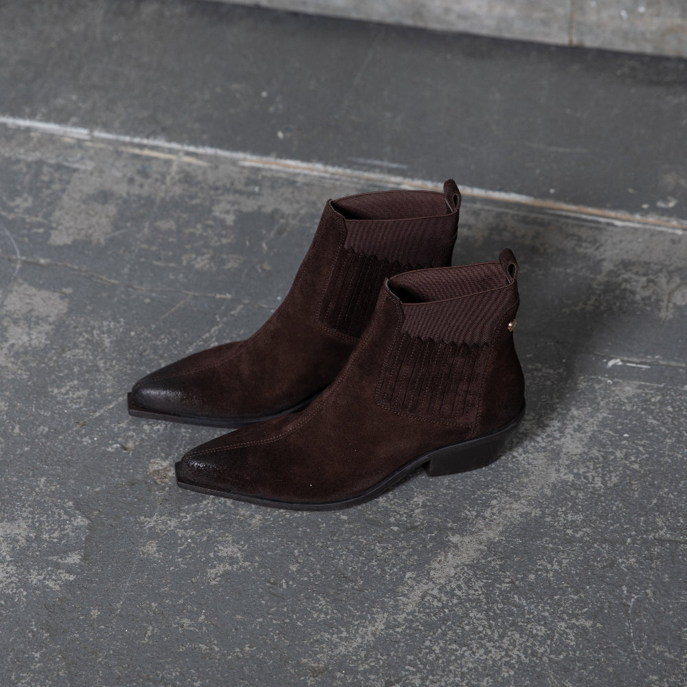 A pair of dark brown suede ankle boots with elastic side panels and pointed toes, displayed on a textured gray concrete floor.