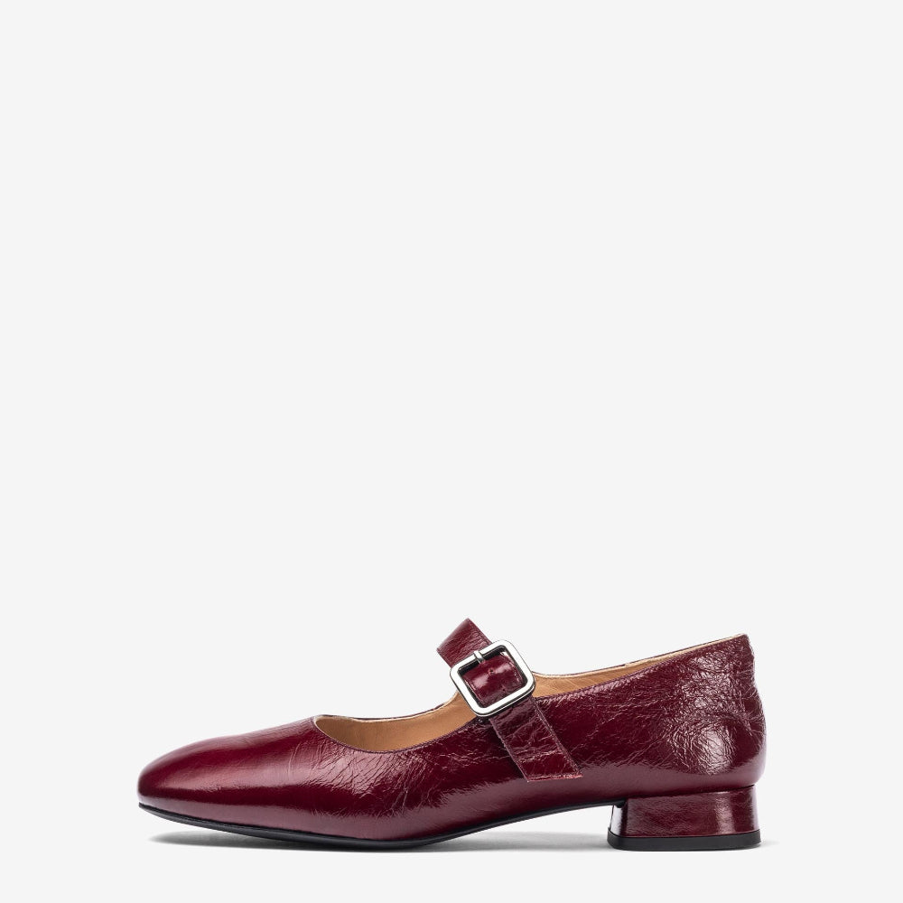 A single women's Mary Jane flat shoe in glossy burgundy textured leather, featuring a low block heel and a decorative buckle strap across the instep, shown against a plain light gray background.