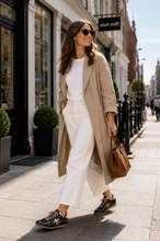 Load image into Gallery viewer, A woman walking on a city street wearing a long beige trench coat, white top and wide-leg white pants, black sneakers with gold star details, and carrying a brown leather handbag.
