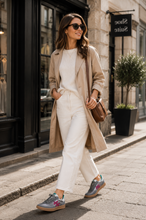 Load image into Gallery viewer, A woman walking on a city sidewalk wearing a beige trench coat, white top and white trousers, gray sneakers with pink and blue accents, and carrying a brown shoulder bag.
