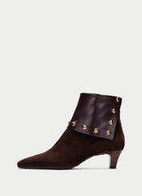 Load image into Gallery viewer, A dark brown suede ankle boot with a pointed toe, a low block heel, and decorative gold-tone metal studs on a matching faux-leather wrap around the collar, shown against a light gray background.
