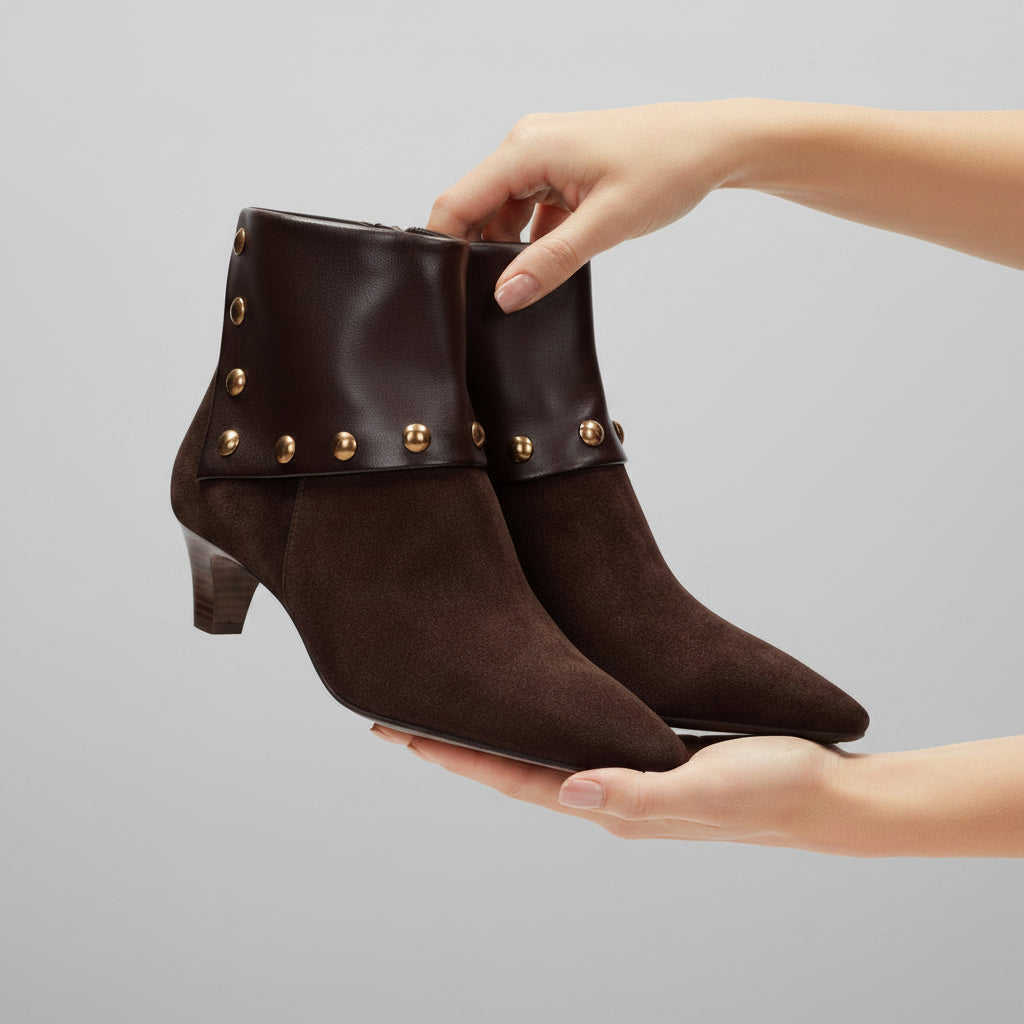 A pair of dark brown suede ankle boots with pointed toes, low block heels, and decorative gold-tone studs along the side, shown against a light gray background.
