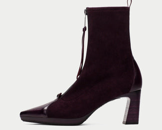 A dark plum purple suede ankle boot with a glossy patent leather toe cap and a block heel, shown from a side profile on a light gray background.