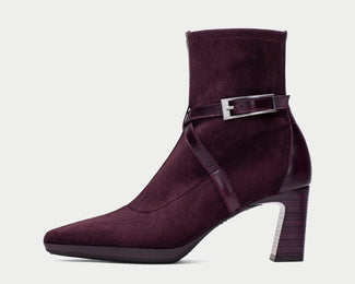 A dark plum purple suede ankle boot with a block heel, pointed toe, and a decorative strap with a silver buckle detail around the ankle, shown against a light gray background.