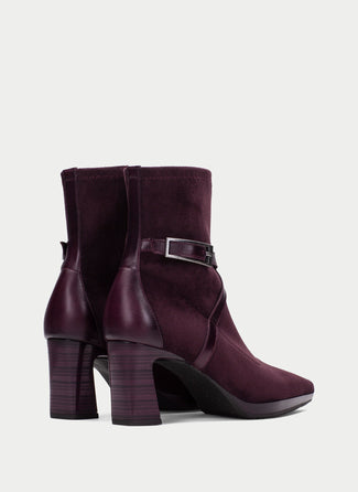 A dark plum purple suede ankle boot with a block heel, pointed toe, and a decorative strap with a silver buckle detail around the ankle, shown against a light gray background.