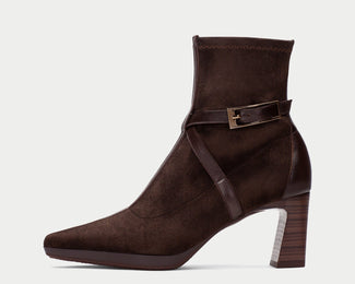 A dark brown suede ankle boot with a pointed toe, a stacked block heel, and a decorative ankle strap with a gold-tone buckle, shown against a light gray background.