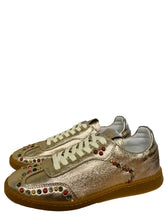 Load image into Gallery viewer, Pair of gold sneakers with red and gold embellishments on a surface near a window.
