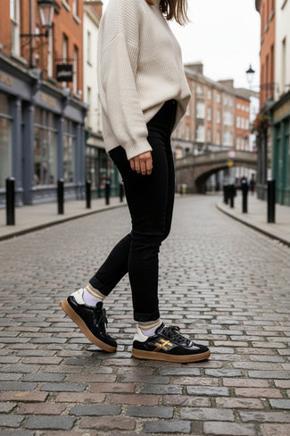 A single black leather sneaker with a prominent gold 'H' logo on the side, a white heel tab, and a beige gum sole, shown standing on a wooden floor against a plain wall.