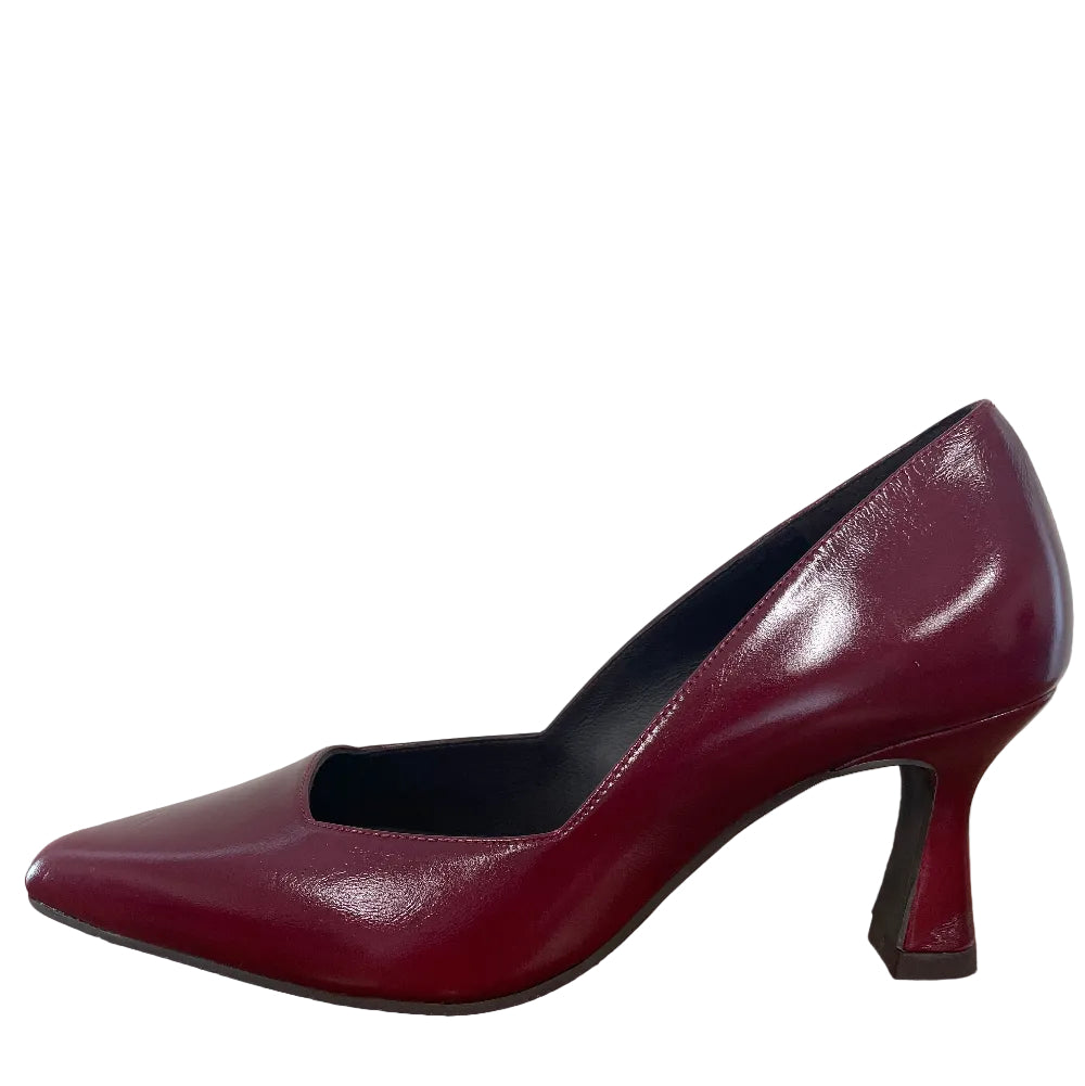 A single burgundy patent leather pump with a pointed toe and block heel, shown standing on a wooden floor against a white wall.