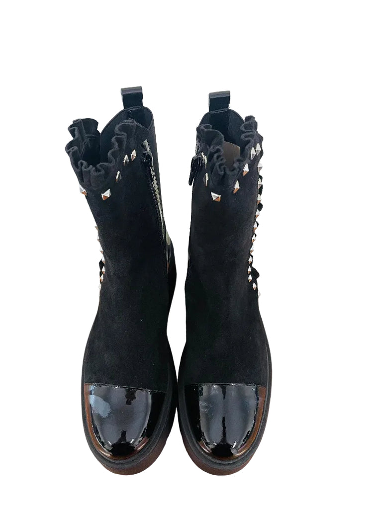 A pair of black ankle boots with ruffled cutout details and shiny patent leather toe caps, displayed on a wooden surface against a white wall.