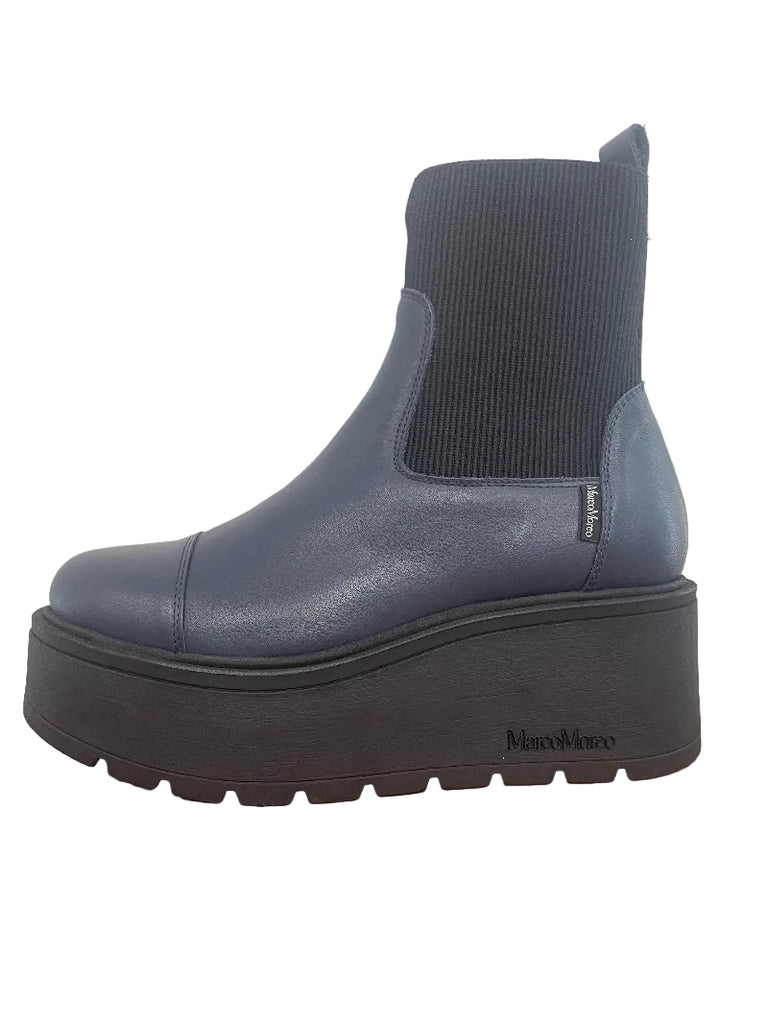 A single platform ankle boot by Marco Moro, featuring a navy blue leather upper, a black ribbed elastic side panel, and a chunky black platform sole, shown on a wooden table against a plain wall.