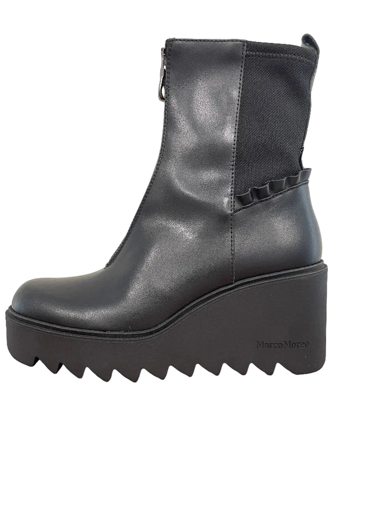 A black leather ankle boot with a chunky platform wedge heel and a scalloped ruffle detail on the side, resting on a wooden table against a plain wall.