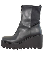 Load image into Gallery viewer, A black leather ankle boot with a chunky platform wedge heel and a scalloped ruffle detail on the side, resting on a wooden table against a plain wall.
