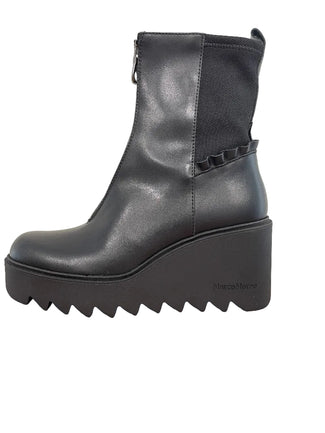 A black leather ankle boot with a chunky platform wedge heel and a scalloped ruffle detail on the side, resting on a wooden table against a plain wall.
