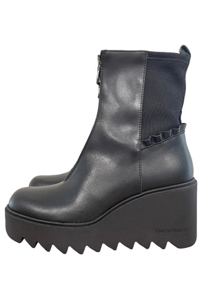 A black leather ankle boot with a chunky platform wedge heel and a scalloped ruffle detail on the side, resting on a wooden table against a plain wall.