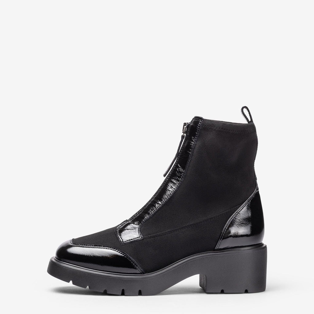 A black ankle boot with a chunky lug sole, glossy patent leather toe cap, and side zipper, shown from a side profile on a light gray background.