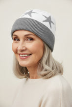 Load image into Gallery viewer, A white knit beanie hat with three gray stars knitted across the top and a ribbed gray cuff, photographed on a white background with a green fern leaf at the upper left.
