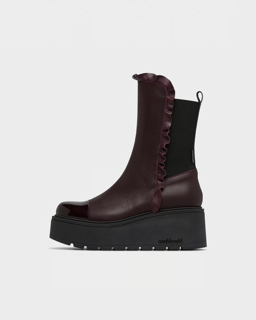 A dark burgundy leather ankle boot with a black elastic side panel, ruffled trim, and a chunky black platform sole, shown from a side profile on a light gray background.