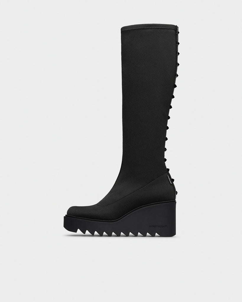 A single black knee-high boot with a chunky wedge platform sole and a row of sharp teeth-like studs running down the outer side, shown in profile against a light gray background.