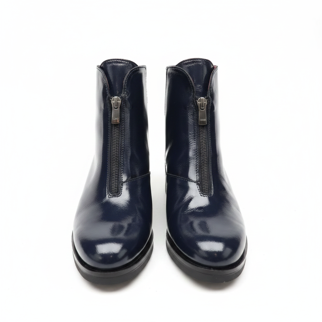 A pair of dark navy blue patent leather ankle boots with front zippers, photographed on a light gray surface against a gray background.