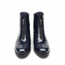 Load image into Gallery viewer, A pair of dark navy blue patent leather ankle boots with front zippers, photographed on a light gray surface against a gray background.
