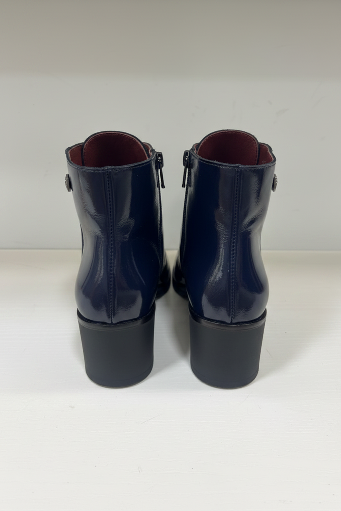 A pair of navy blue patent leather ankle boots with low block heels, shown from the back on a white surface against a white wall.