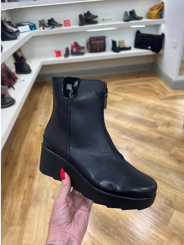 A black leather ankle boot with a chunky platform sole and side zipper, held by a hand with red nail polish in a shoe store.