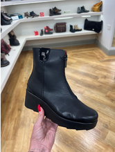 Load image into Gallery viewer, A black leather ankle boot with a chunky platform sole and side zipper, held by a hand with red nail polish in a shoe store.
