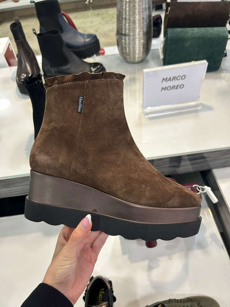 A hand holds up a brown suede ankle boot with a chunky platform sole in a retail store setting. Other boots and a sign reading 'MARCO MOREO' are visible in the background.