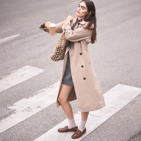 Woman in a trench coat crossing a street with a coffee cup and bag.